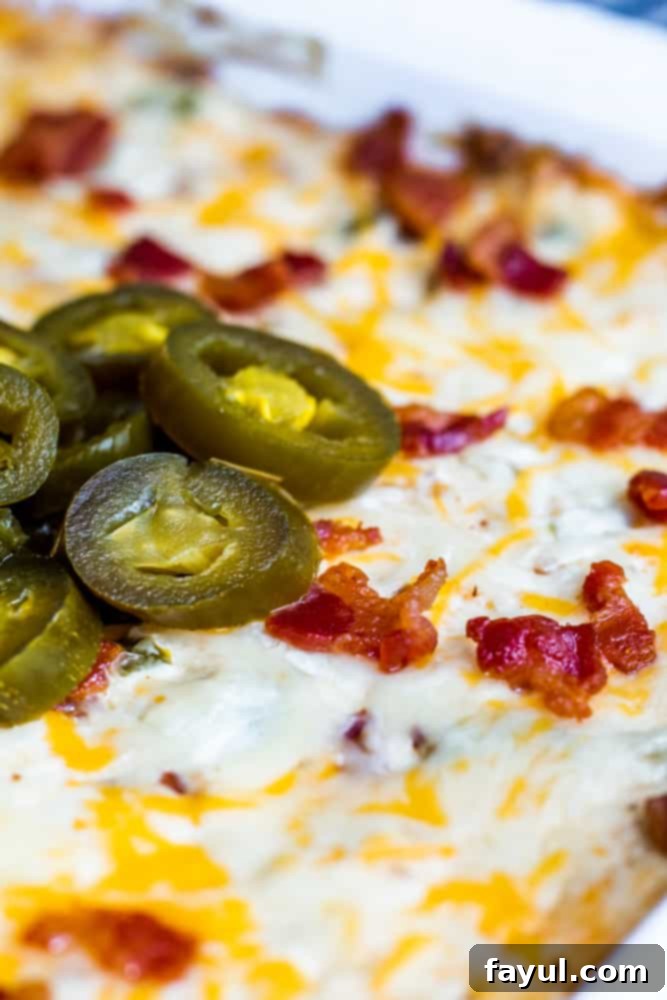 This Baked Bacon Jalapeno Popper Dip with Bacon is the best, easy appetizer recipe! Perfect for game days, parties, holidays or just for the family and ready in only 30 minutes. Pair with tortilla chips, Ritz Crackers or Veggie Sticks for the perfect finish. Simple step by step instructions for crockpot and oven, recipe alterations, and video included! #dip #cheese #JalapenoDip #Spicy #bacon #appetizer #recipe #gamedays #Thanksgiving #Christmas Close up shot of jalapenos on top of dip in a white dish.