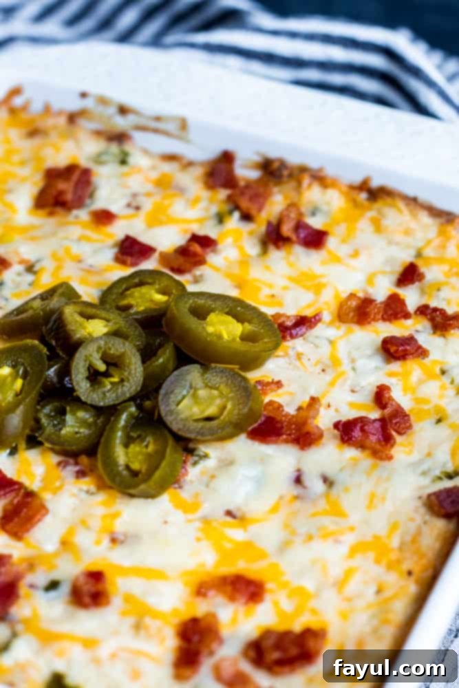 Holy Bacon and Cheese! This Baked Bacon Jalapeno Popper Dip with Bacon is the best, easy appetizer recipe! Perfect for game days, parties, holidays or just for the family and ready in only 30 minutes. Pair with tortilla chips, Ritz Crackers or Veggie Sticks for the perfect finish. Simple recipe step by step instructions for crockpot and oven, recipe alterations, and video included! #dip #cheese #JalapenoPopperDip #Spicy #bacon #appetizer #recipe #gamedays Shot of dip recipe in white dish with blue and white towel in the background.