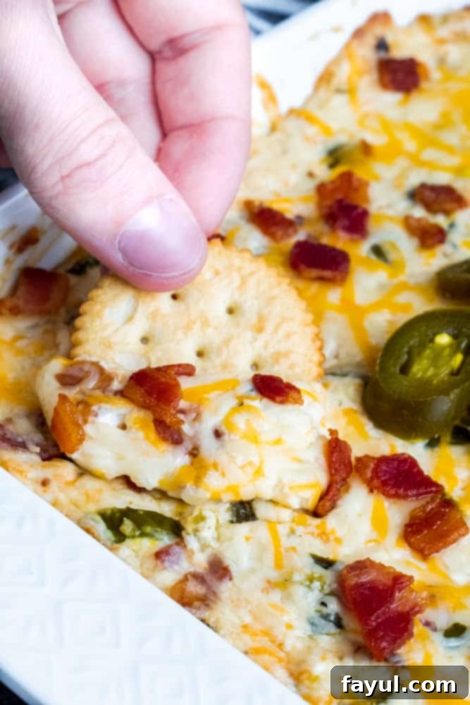 This Baked Bacon Jalapeno Popper Dip with Bacon is the best, easy appetizer recipe! Perfect for game days, parties, holidays or just for the family and ready in only 30 minutes. Pair with tortilla chips, Ritz Crackers or Veggie Sticks for the perfect finish. Simple step by step instructions for crockpot and oven, recipe alterations (freezer friendly, prep ahead and vegetarian), and video included! #dip #cheese #JalapenoDip #Spicy #bacon #appetizer #recipe #gamedays Cracker being dipped into JDip with Bacon y a hand in a white dish.