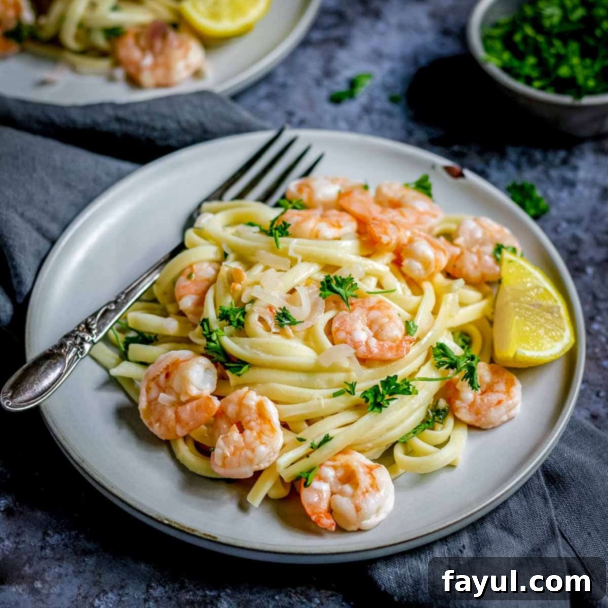 Side shot of a perfectly plated serving of shrimp scampi garnished with fresh parsley on a modern grey plate, with a fork ready on a black and grey surface. A bowl of fresh herbs is blurred in the background, hinting at the fresh ingredients.