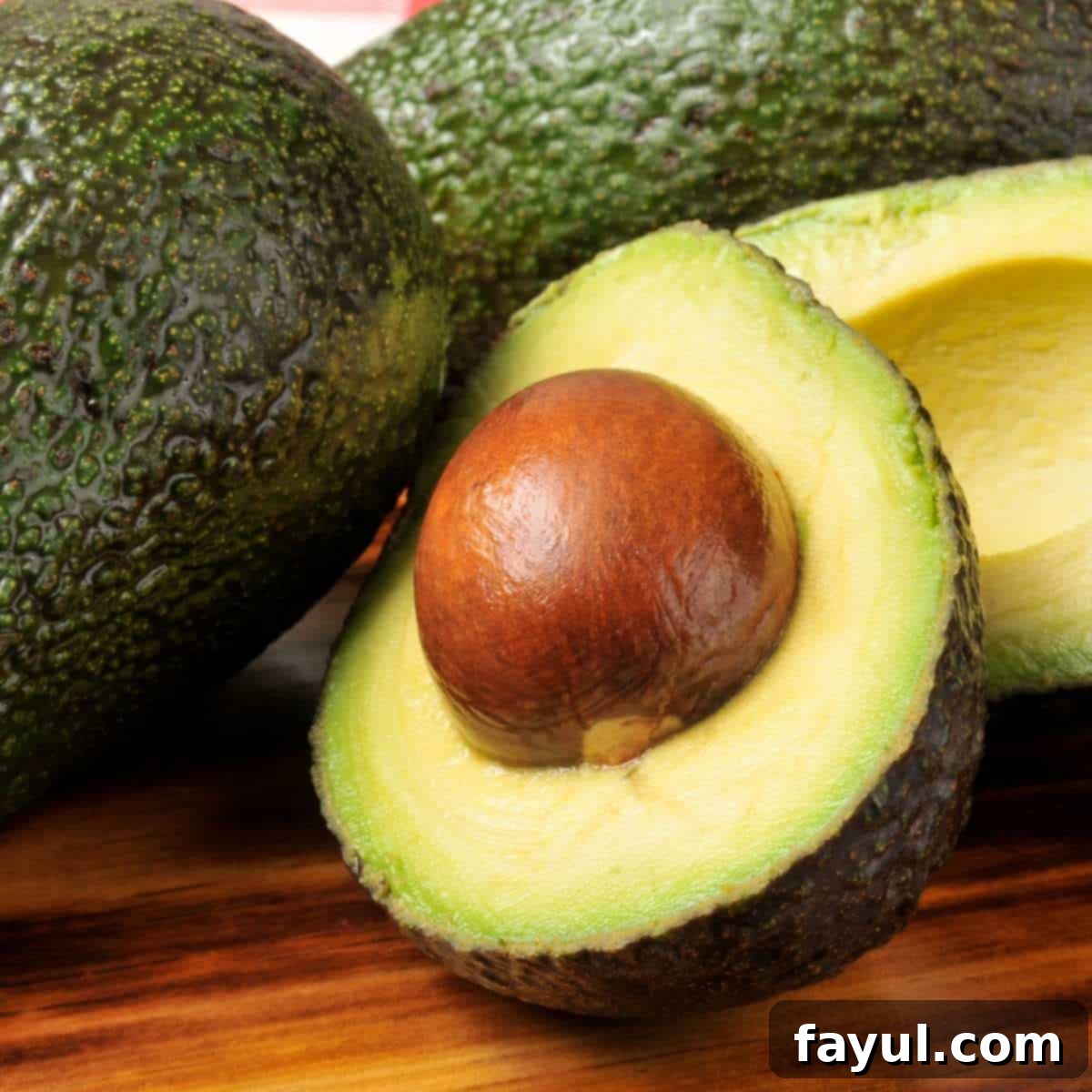 Avocado Flavor Demystified 3 Hass avocado half with pit still intact, on a wooden surface with more avocados in the background.