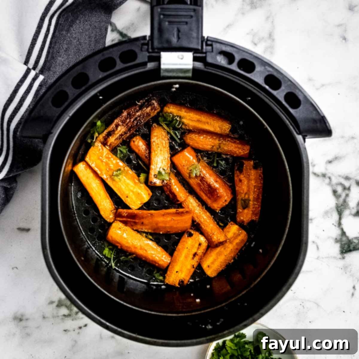 Overhead shot of roasted carrots with herbs in the air fryer on a marble countertop with a black and white striped cloth on the side.
