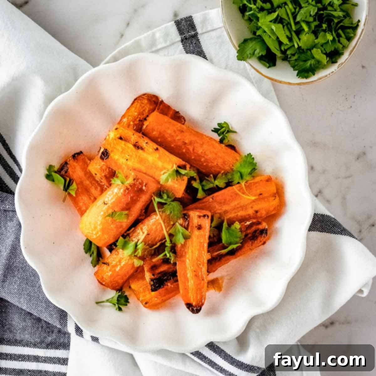 Overhead shot of roasted carrots garnished with herbs on a white plate on a grey and white cloth with a small bowl of herbs on the side.