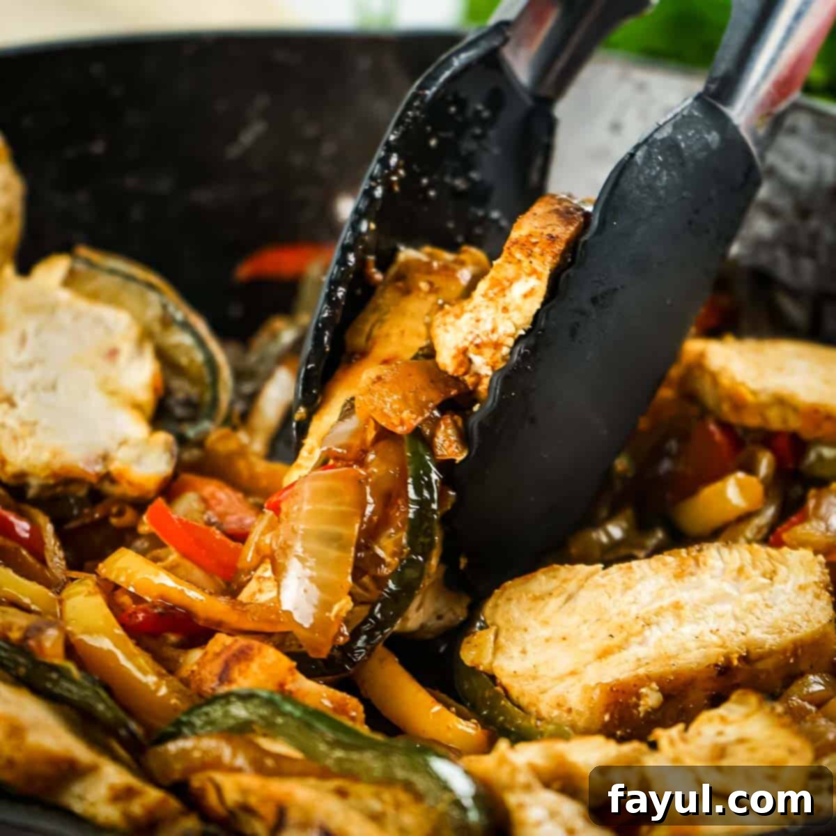 Side close-up shot of black tongs picking up some perfectly cooked chicken fajita filling (chicken, peppers, and onions) directly out of a black cast iron skillet, showcasing the delicious sizzle.