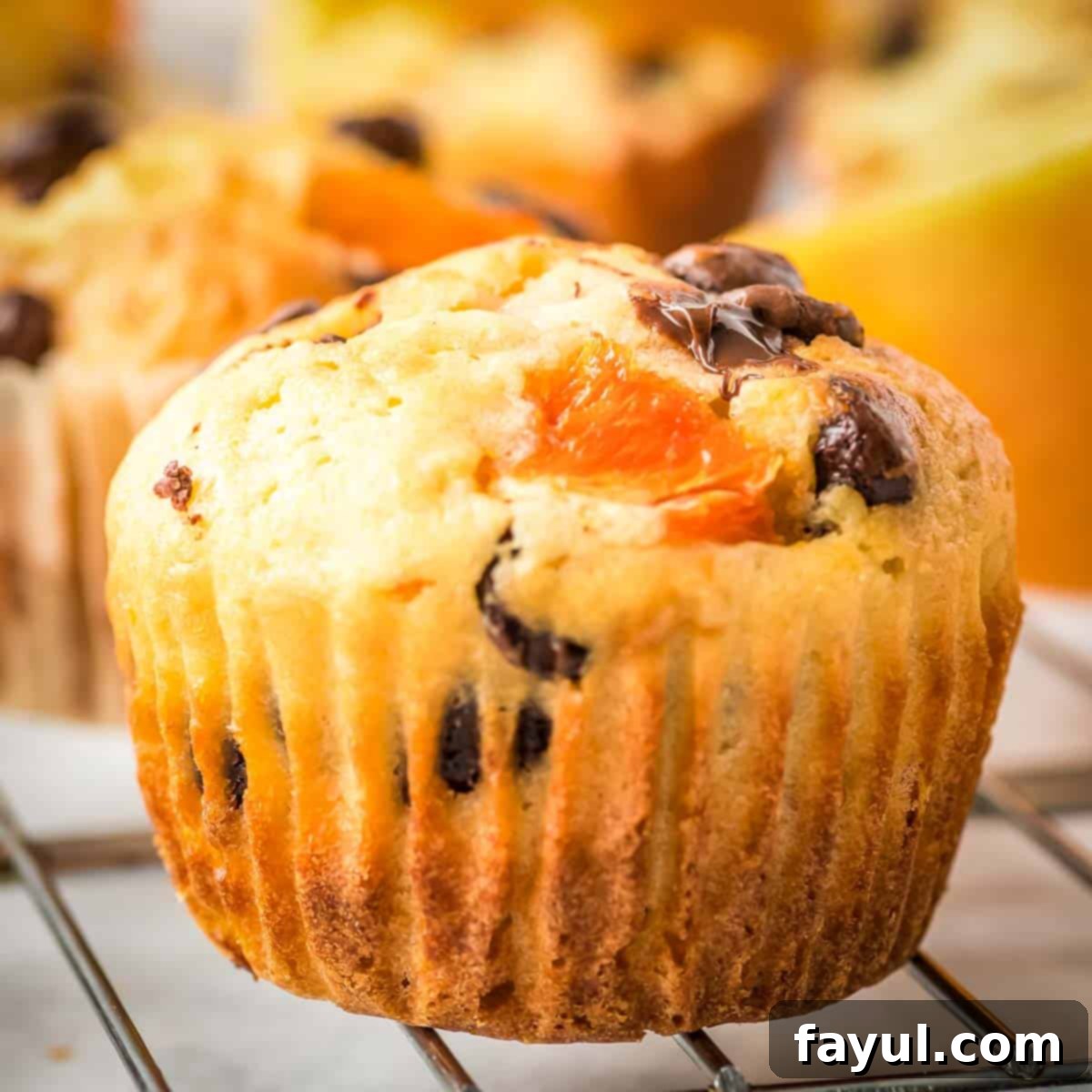 Close up side shot of one orange chocolate muffin on a cooling rack with more muffins blurred in the background, showcasing its fluffy texture.
