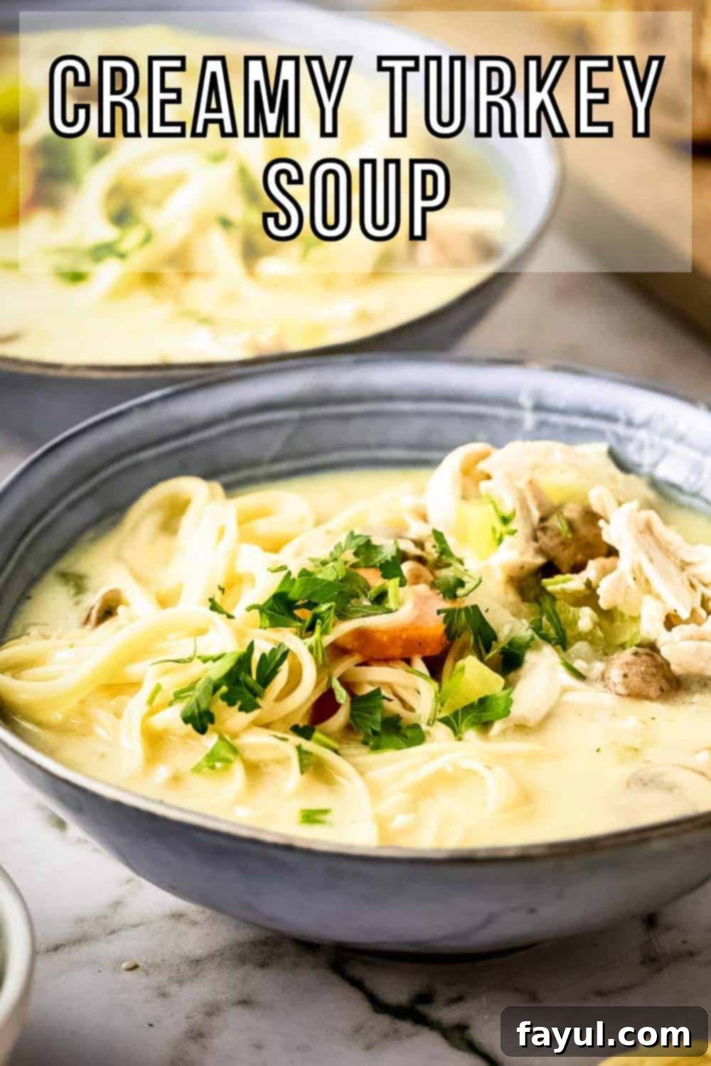 Side shot of two grey bowls with creamy turkey noodle soup garnished with fresh herbs on a marble countertop with a wooden cutting board and sliced bread in the background.