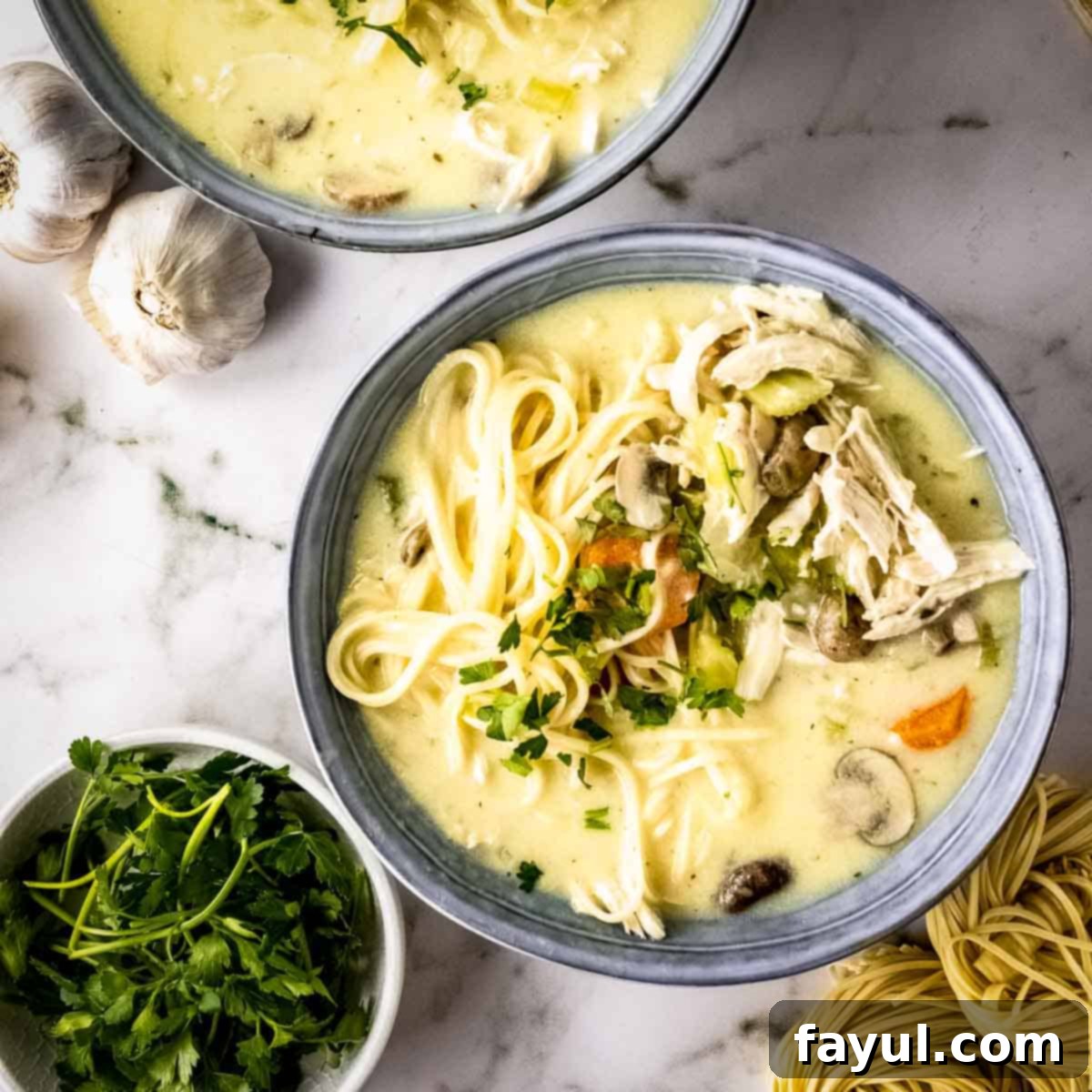 Overhead shot of two grey bowls with creamy turkey noodle soup garnished with fresh herbs on a marble countertop, with a bowl of parsley, garlic cloves, and uncooked egg noodles on the side.