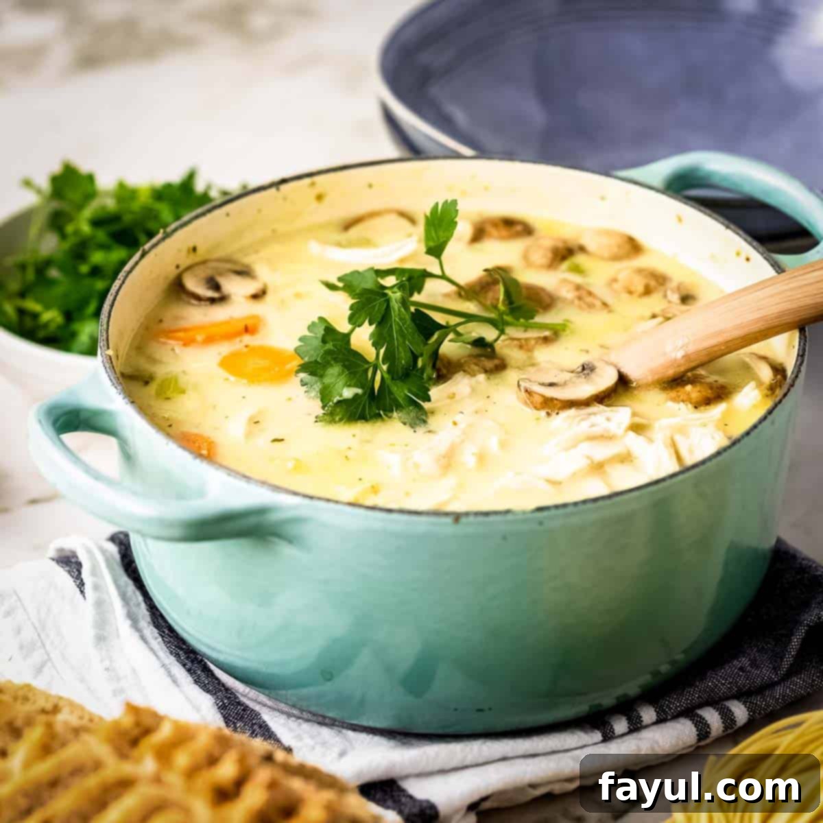Side shot of a large teal colored pot filled with turkey noodle soup garnished with fresh parsley on a dark blue and white cloth with a wooden spoon leaning on the edge and a bowl of parsley in the background.