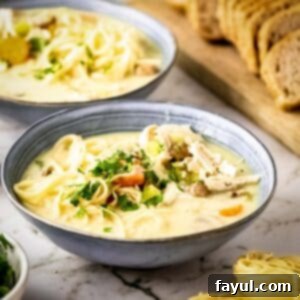 Side shot of two grey bowls with turkey noodle soup garnished with fresh herbs on a marble countertop with a wooden cutting board and sliced bread in the background.