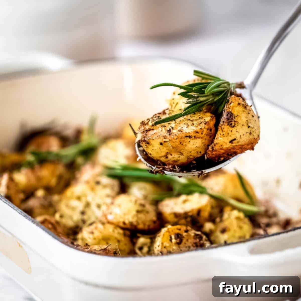 Side shot of a spoon picking up a few potatoes from a white pan garnished with fresh rosemary.