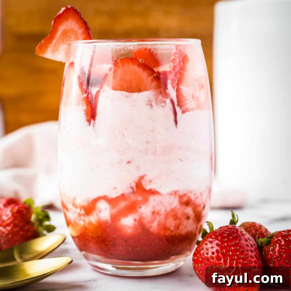 Side shot of a glass tumbler with strawberry mousse and sliced strawberries on a. marble table with gold spoons and strawberries on the side.