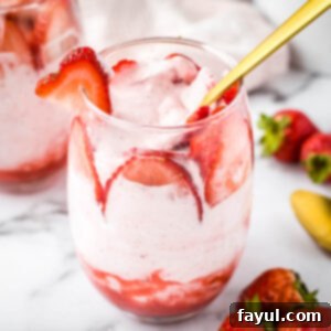 Side close up shot of a glass tumbler with strawberry mousse and sliced strawberries with a gold spoon on a marble table.