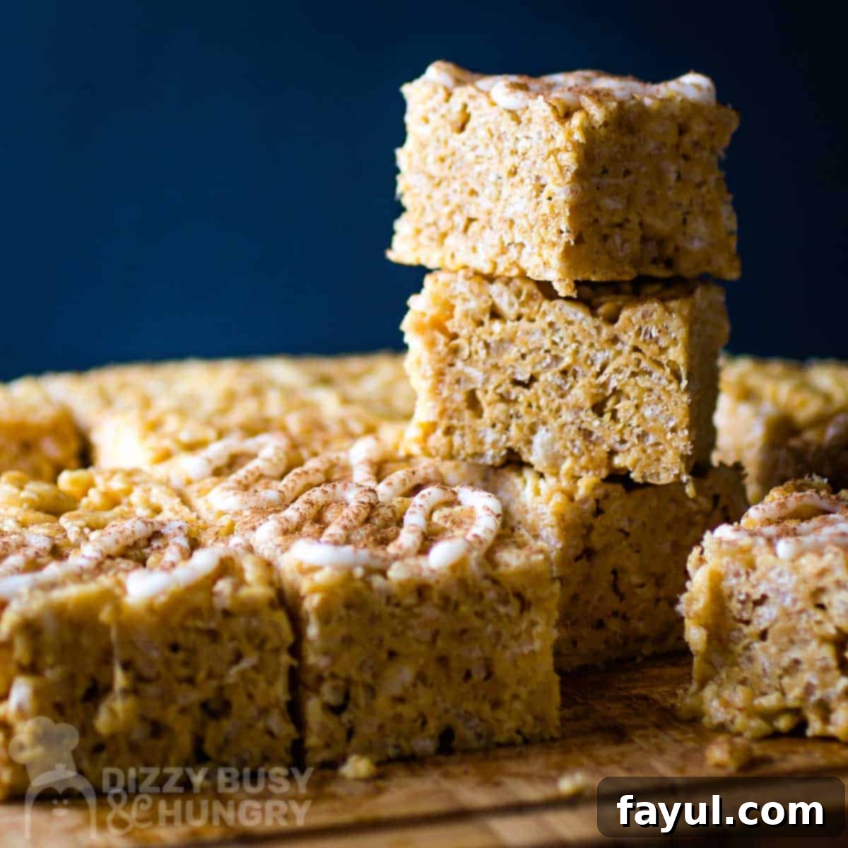 Side shot of multiple Rice Krispies with an icing drizzle stacked on each other on a wooden cutting board, ready to be enjoyed.