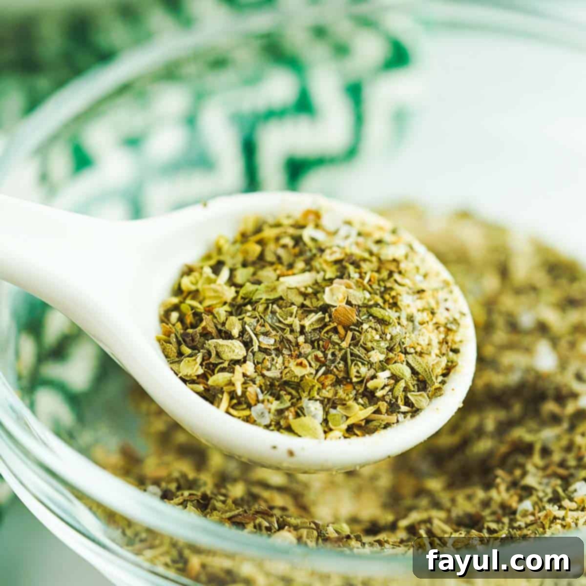 Side shot of a white spoon holding up some greek spice mix over a small clear bowl full of the mix.