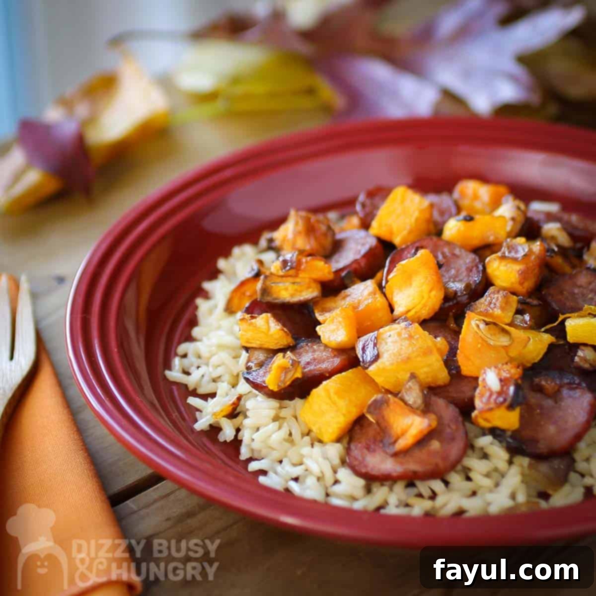 Side close up shot of sausage and butternut squash on a bed of rice on a red plate with an orange napkin and fork on the side.