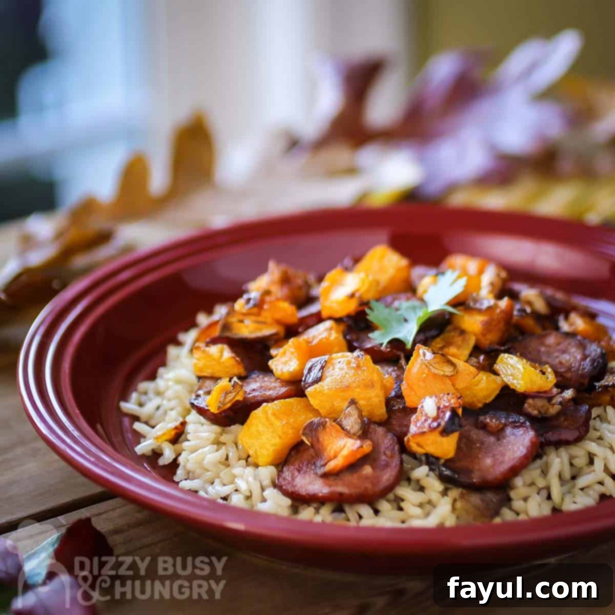 Side shot of sausage and butternut squash on a bed of rice on a red plate with fake leaves in the background.
