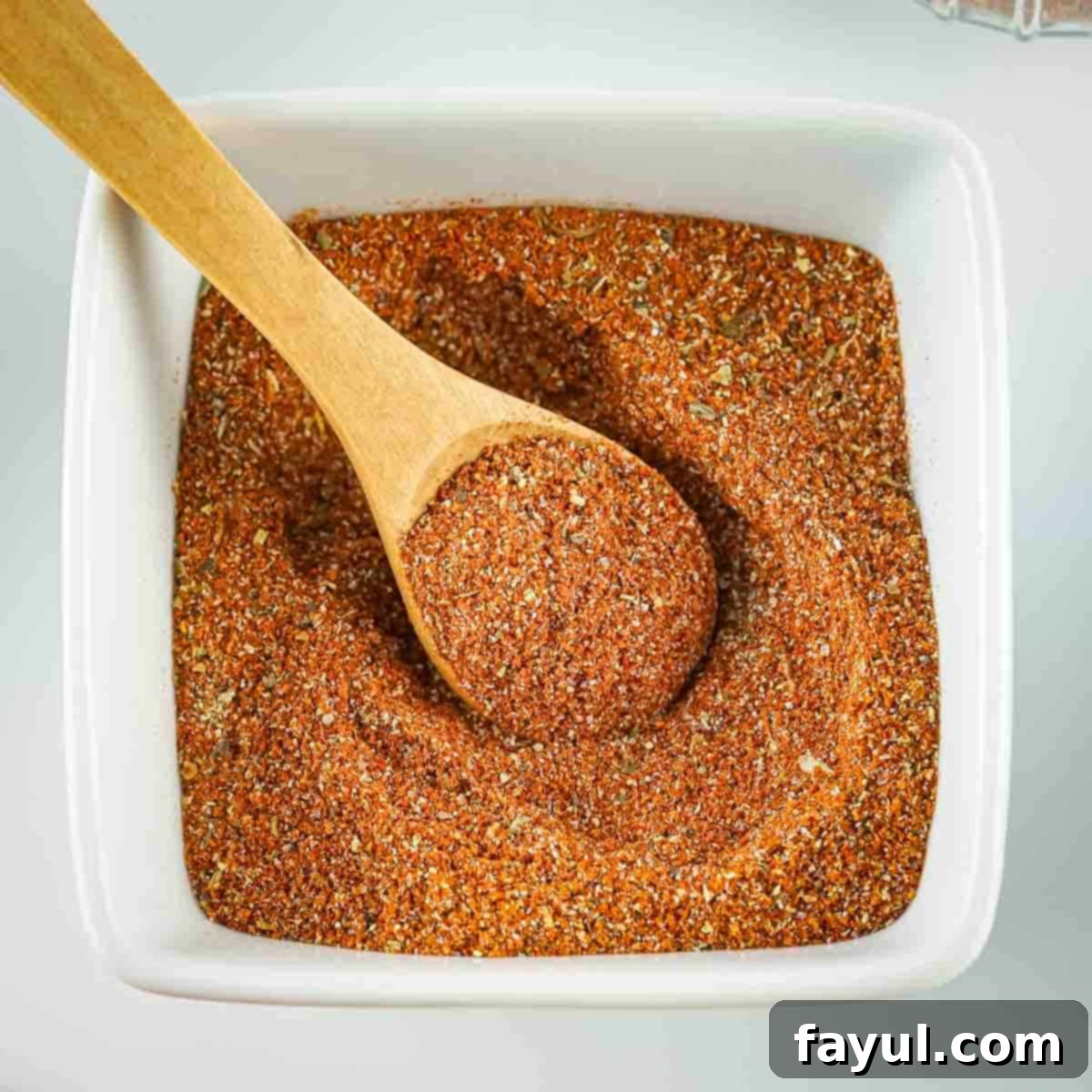 Overhead shot of blackened seasoning in a square white bowl with a wooden spoon scooping some seasoning out.
