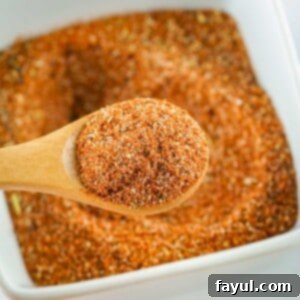 Overhead close up shot of a wooden spoon holding a scoop of seasoning over a white bowl full of blackened seasoning on a white surface.