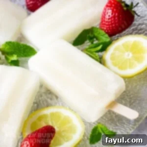 Side shot of lemonade popsicles on a glass plate with mint, lemon slices, and strawberries scattered around.