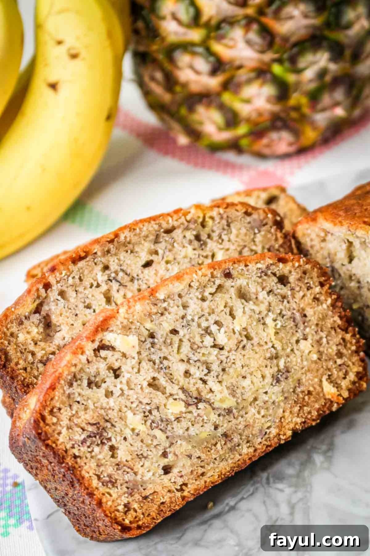 Side shot of slices of pineapple banana bread on a marble plate with bananas and a pineapple in the background.