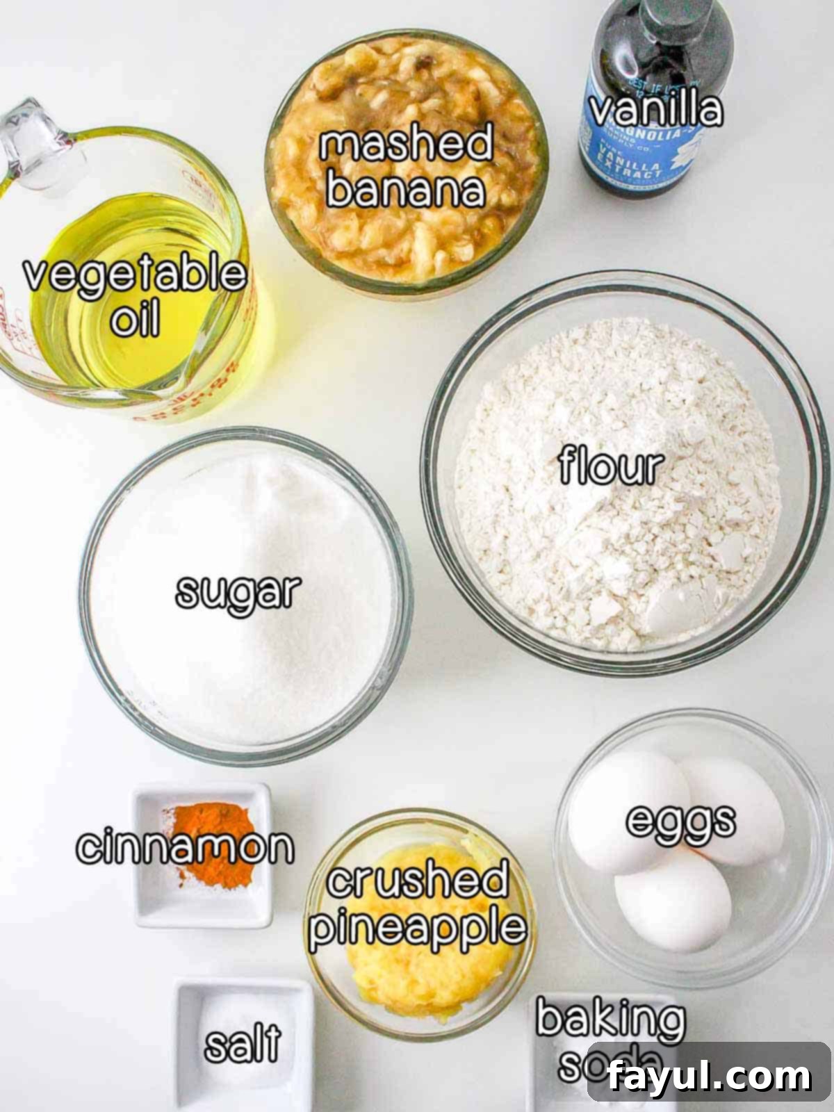Overhead shot of ingredients - vanilla, mashed banana, vegetable oil, sugar, flour, cinnamon, crushed pineapple, eggs, salt, and baking soda.