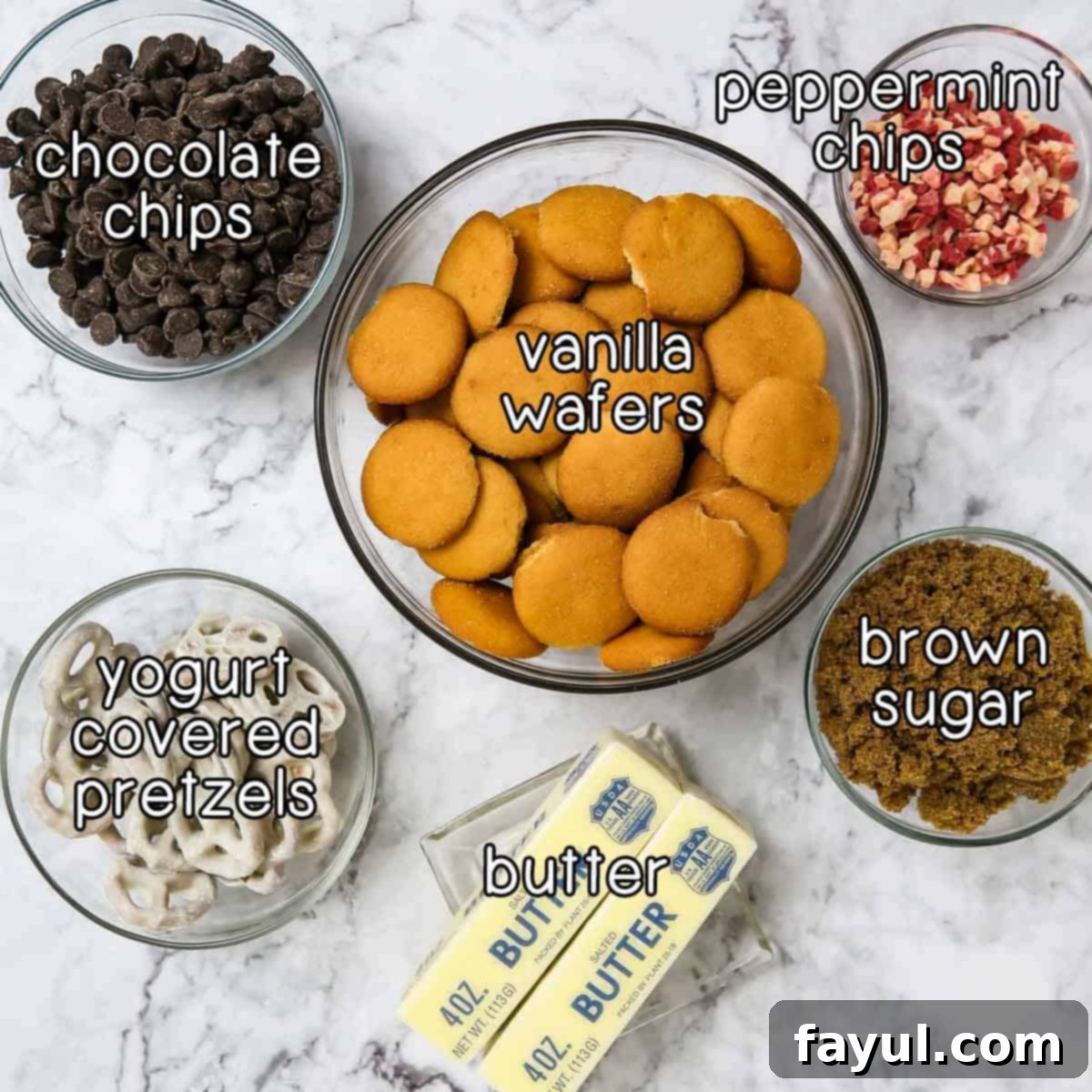 Overhead shot of Christmas bark ingredients including vanilla wafers, peppermint chips, brown sugar, butter, yogurt covered pretzels, and chocolate chips, neatly arranged on a rustic surface.