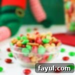 Side view of Christmas snack mix in a white bowl with M&Ms scattered around on a white table, with Christmas decorations in the background.