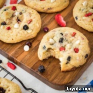 Side shot of multiple red, white, and blue cream cheese cookies, one with a bite taken out, arranged on a rustic wooden cutting board with freeze-dried fruits scattered around for decoration. The cookies are soft, chewy, and studded with colorful fruit pieces and white chocolate.
