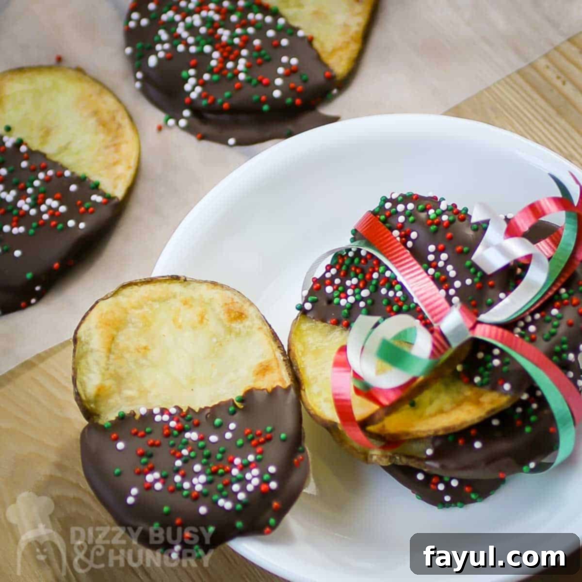 Overhead close-up shot of multiple chocolate covered potato chips artfully arranged with Christmas ribbons on a white plate, with more in the background on a rustic wooden surface.