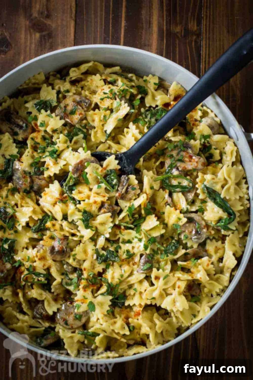 Overhead shot of creamy spinach and mushroom pasta in a large pot with a rubber spoon, ready to serve on a wooden table. The pasta is perfectly coated in a rich, creamy sauce with visible spinach and sliced mushrooms.