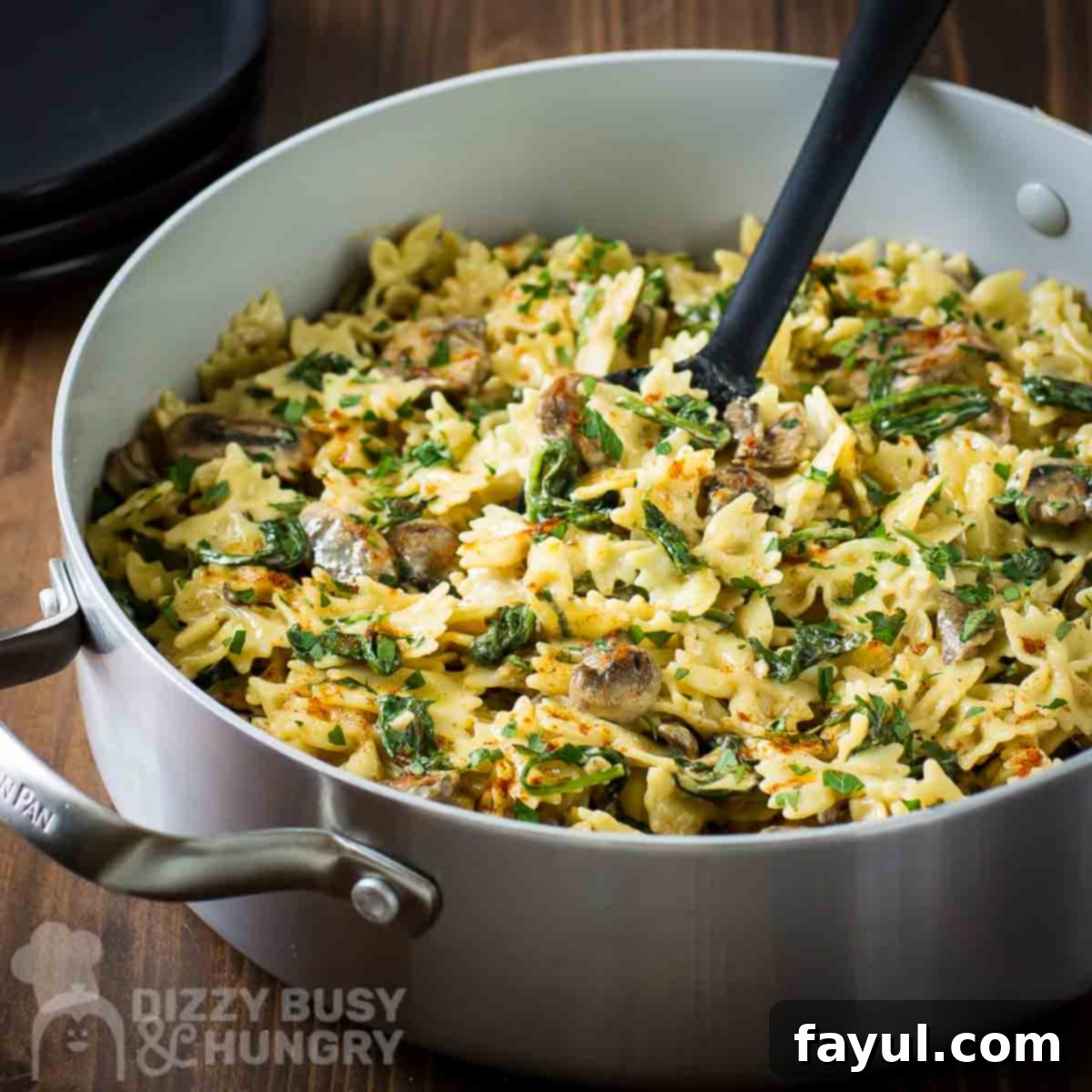 Side angled shot of creamy spinach and mushroom pasta in a silver pan with a black spoon on a rustic wooden surface, showcasing the rich texture and appealing presentation of the dish.