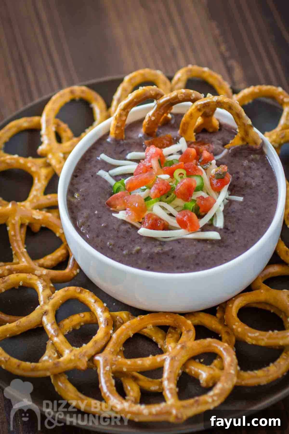 Side angled shot of black bean dip garnished with diced tomatoes, herbs, and cheese in a white bowl on a black plate with pretzels on the side all on a wooden surface.