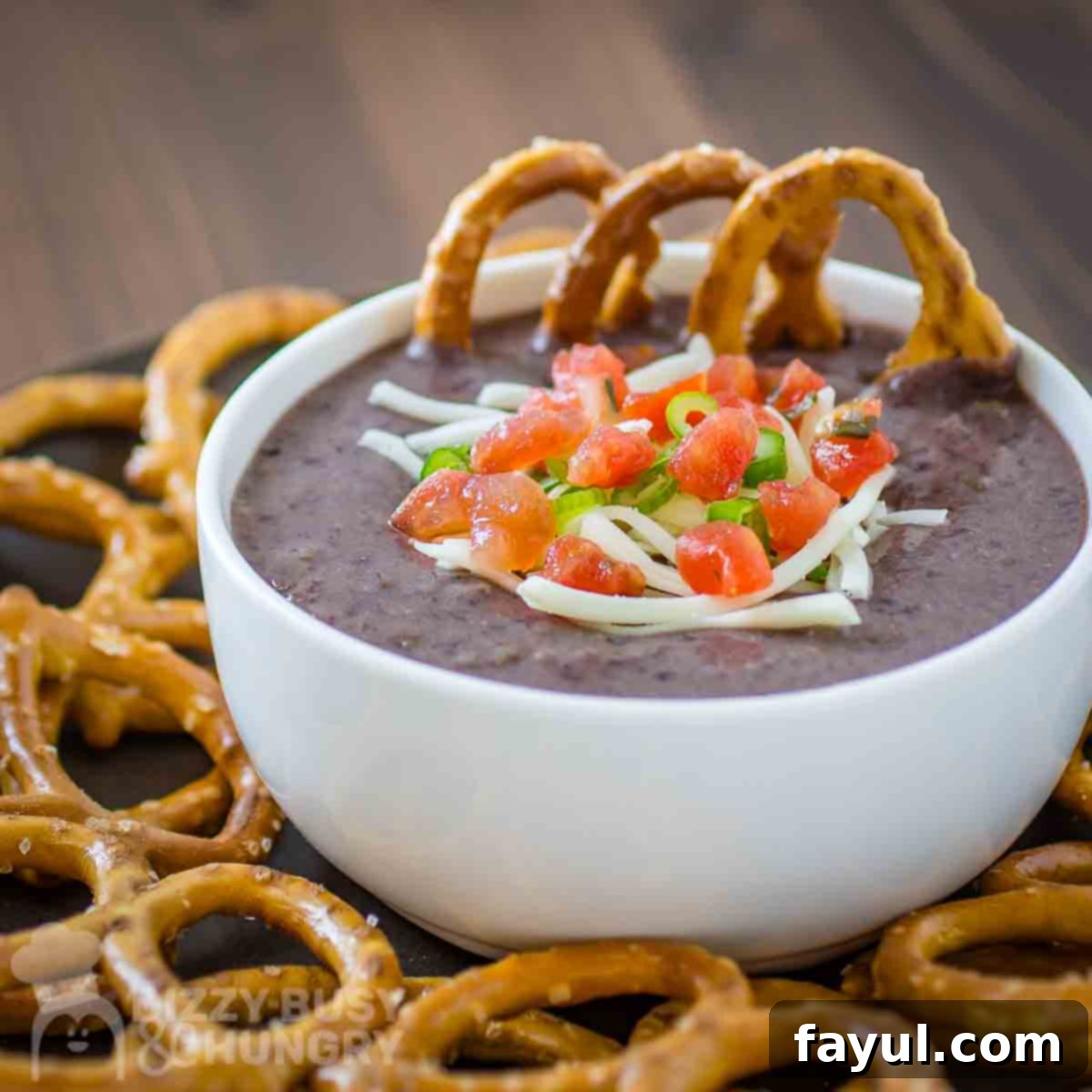 Side shot of black bean dip garnished with diced tomatoes, herbs, and cheese in a white bowl on a black plate with pretzels on the side.