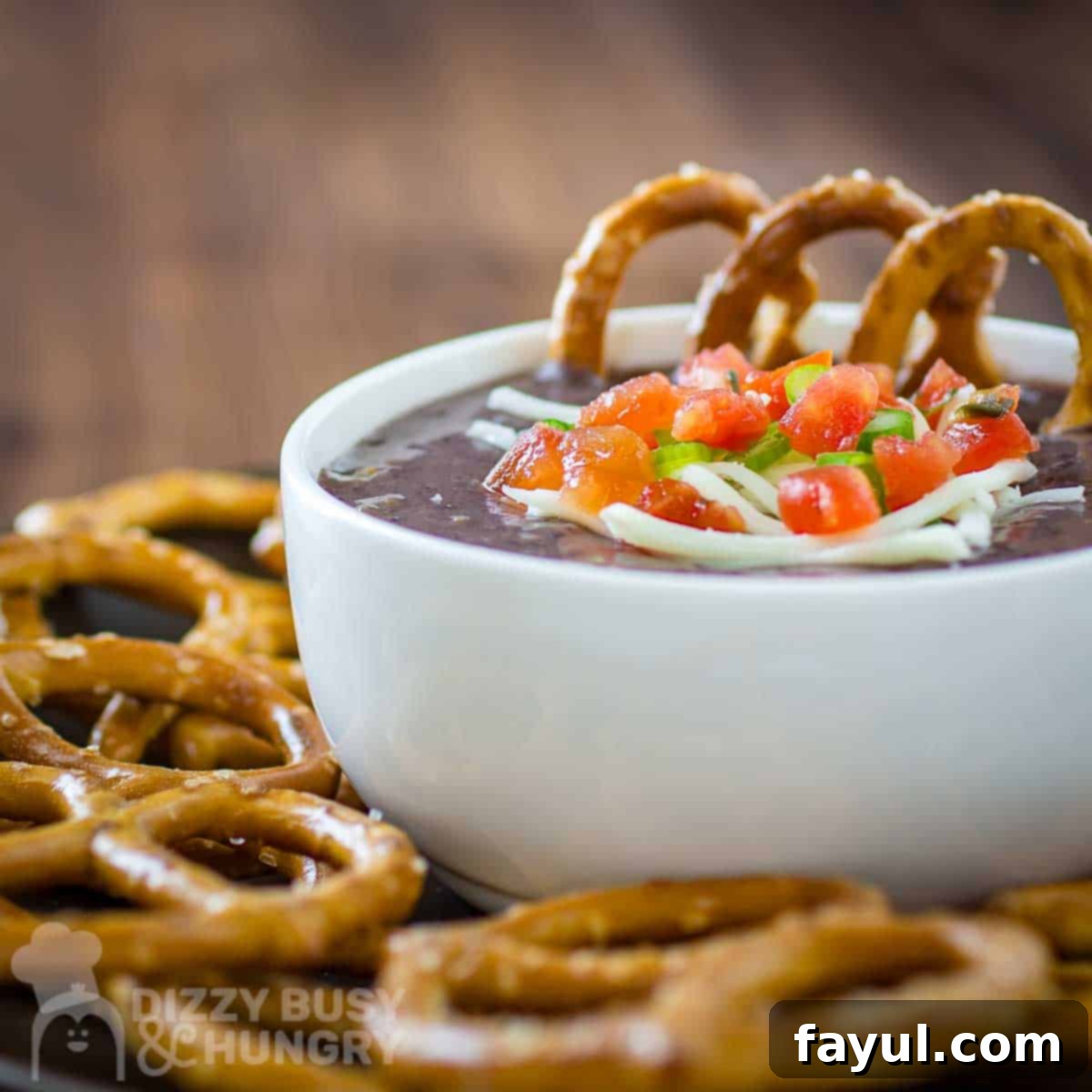 Side shot of black bean dip garnished with diced tomatoes, herbs, and cheese in a white bowl on a black plate with pretzels on the side.