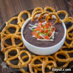 Side angled shot of black bean dip garnished with diced tomatoes, herbs, and cheese in a white bowl on a black plate with pretzels on the side all on a wooden surface.