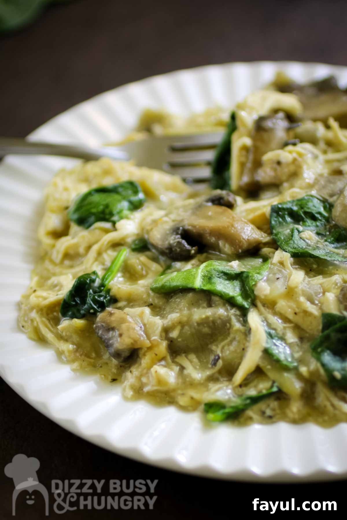 Side close up shot of creamy slow cooker chicken and mushrooms with artichoke hearts on a white plate with a fork on the side.