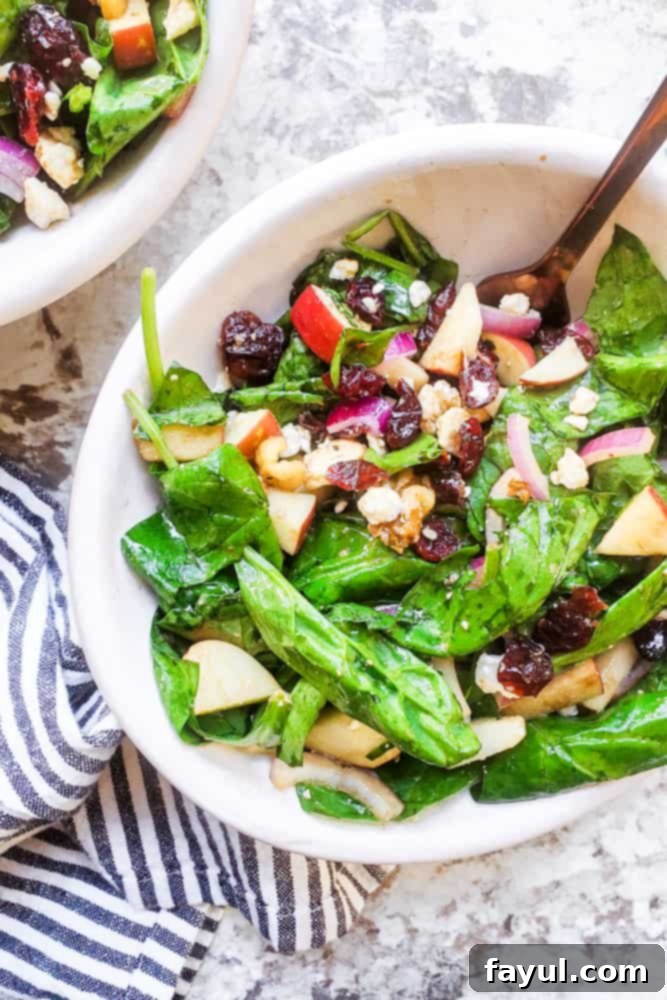 The BEST Healthy Spinach Apple Salad with Homemade Honey Balsamic Vinaigrette. This recipe is perfect for holidays, a side dish or main course. This vegetarian salad is packed with dried cranberries, feta, red onion, apples, and walnuts for the perfect combo. Video included! #salad #healthy #recipe #Food #vegetarianfood #vegetarian Salad in a white bowl with gold fork in it on a white counter.