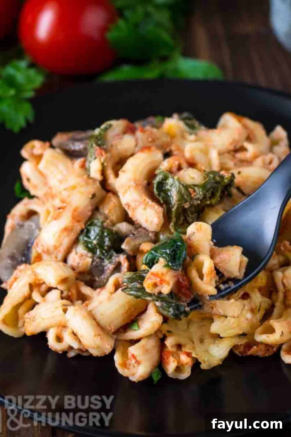 Side close up shot of a fork holding a piece of pasta bake in front of a black plate and a tomato and spinach in the background.