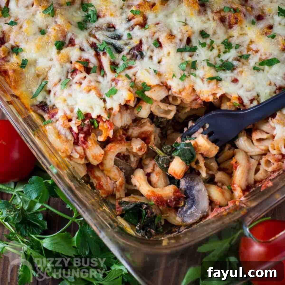 Close up overhead shot of a fork taking a scoop of pasta bake from a casserole dish with herbs and tomatoes on the side.