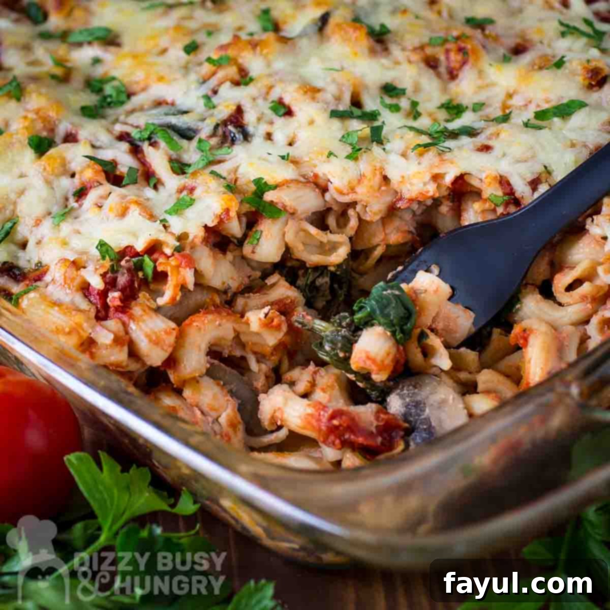 Close up side shot of a fork taking a scoop of pasta bake from a casserole dish with herbs and tomatoes on the side.