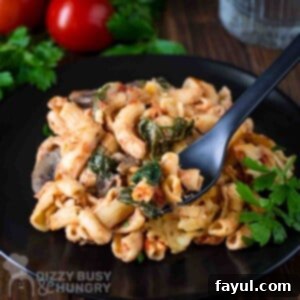 Side close up shot of a fork holding a piece of pasta bake in front of a black plate with it and a tomato and spinach in the background.