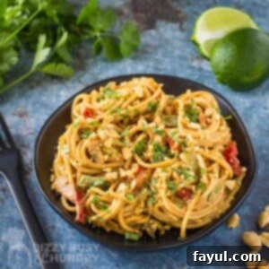 Side angled shot of Thai noodles topped with cilantro and chopped peanuts on a black plate on a blue surface with cilantro and lime wedges in the background.