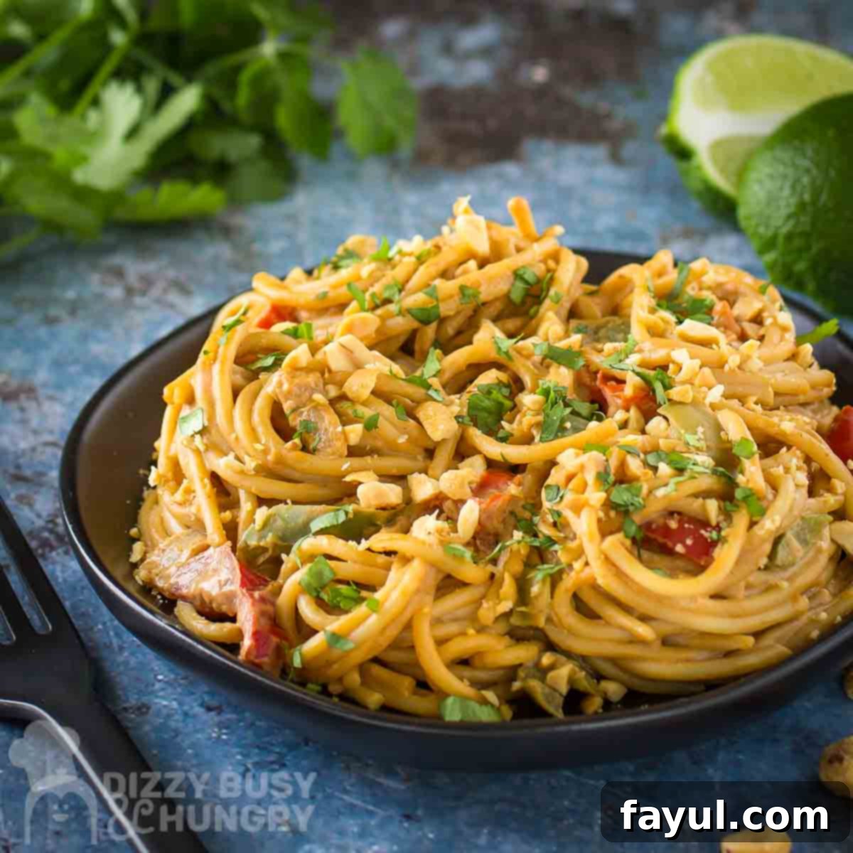 A side close-up shot of a serving of One Pot Spicy Thai Noodles, generously topped with fresh cilantro and chopped peanuts, presented on a black plate. In the softly blurred background, more cilantro and bright lime wedges add a fresh touch to the blue surface.