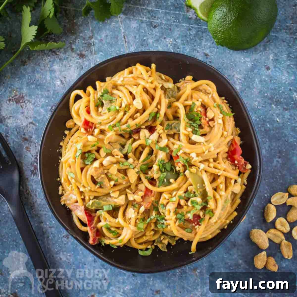 An inviting overhead shot of One Pot Spicy Thai Noodles, beautifully garnished with fresh cilantro and crushed peanuts, served on a black plate. The blue background is softly blurred with cilantro, lime wedges, and whole peanuts, creating a cohesive and appetizing scene.