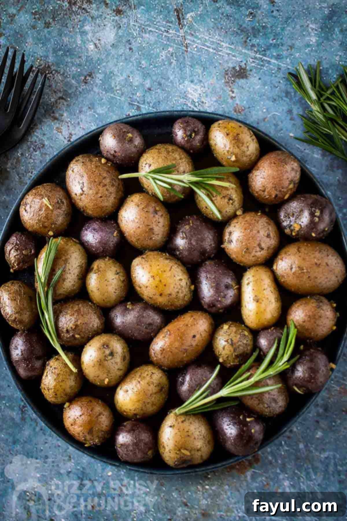 Overhead shot of roasted potatoes garnished with fresh rosemary on a black plate with black silverware and rosemary on the side.