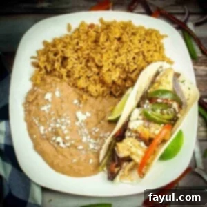 Overhead shot of chicken fajita sprinkled with cotija cheese next to sides of rice and refried beans on a white square plate.