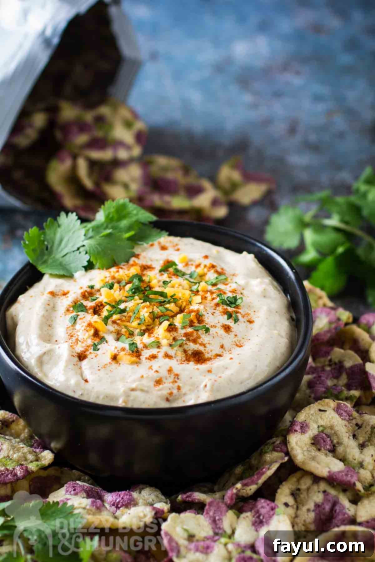 Side angled shot of cheddar cheese dip garnished with herbs and cayenne in a black bowl surrounded by multicolored rice chips.