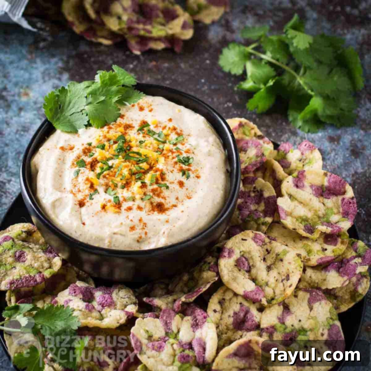 Side angled shot of cheddar cheese dip garnished with herbs and cayenne in a black bowl surrounded by multicolored rice chips.