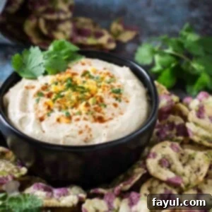 Side angled shot of cheddar cheese dip garnished with herbs and cayenne in a black bowl surrounded by multicolored rice chips.