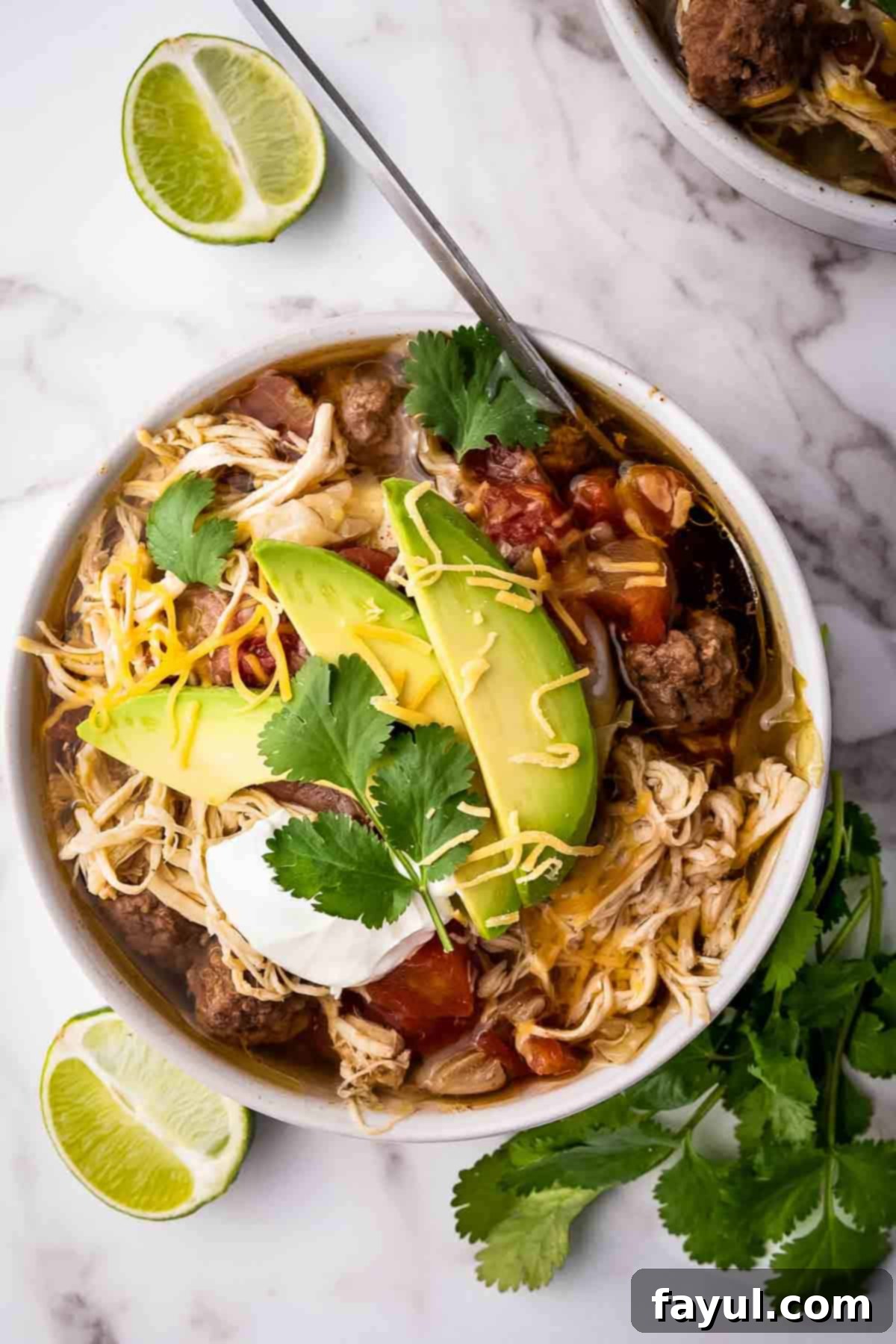 Overhead shot of chicken taco soup with slices of avocado, fresh cilantro, shredded cheese, and sour cream on top all in a white bowl with a lime wedge and fresh cilantro on the side.